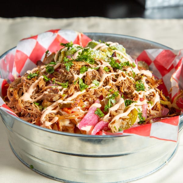 Pulled pork nachos with sauce, cilantro, and pickled onions in a metal bucket with checkered paper.