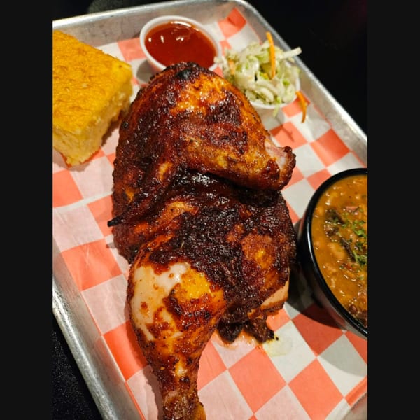 Two pieces of BBQ chicken with cornbread, coleslaw, baked beans, and ketchup on a tray.
