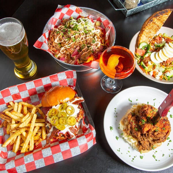 A table spread of American dishes: nachos, a burger with fries, fried chicken, a salad, and drinks.