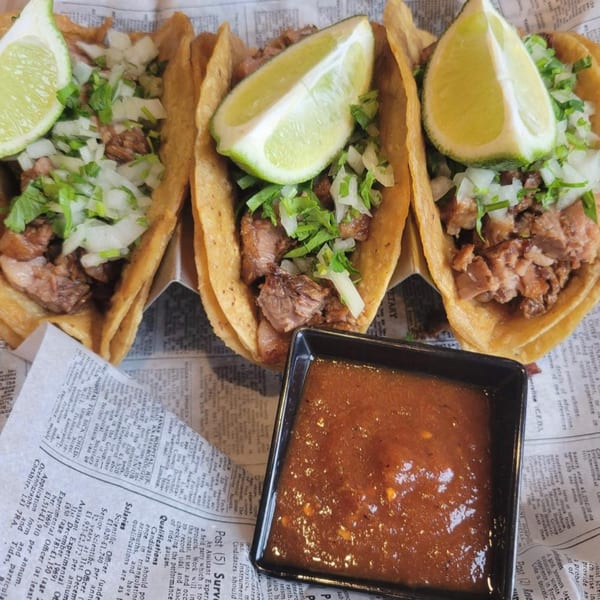 Three American street tacos with meat, onions, cilantro, and lime wedges, served with a side of salsa.