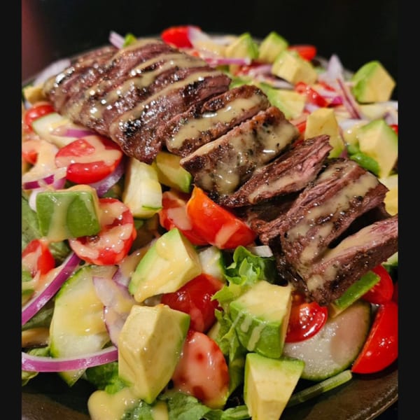 Grilled steak slices with dressing on a fresh salad of avocado, tomatoes, red onion, and lettuce.