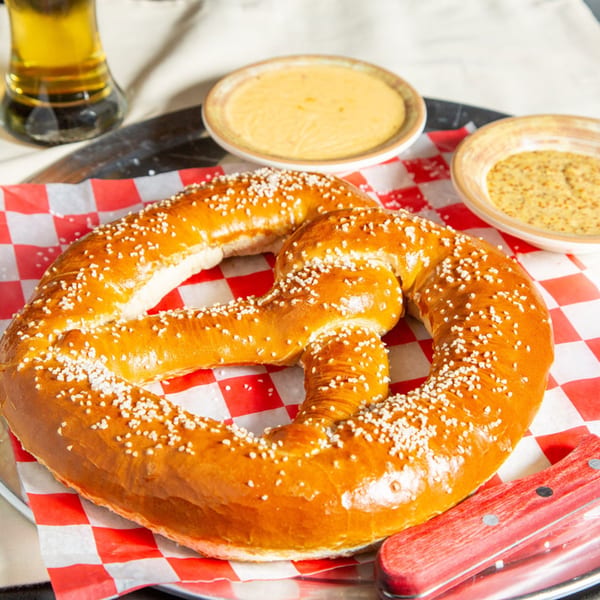 A large, salted pretzel on red and white checkered paper, with two dipping sauces and a beer.