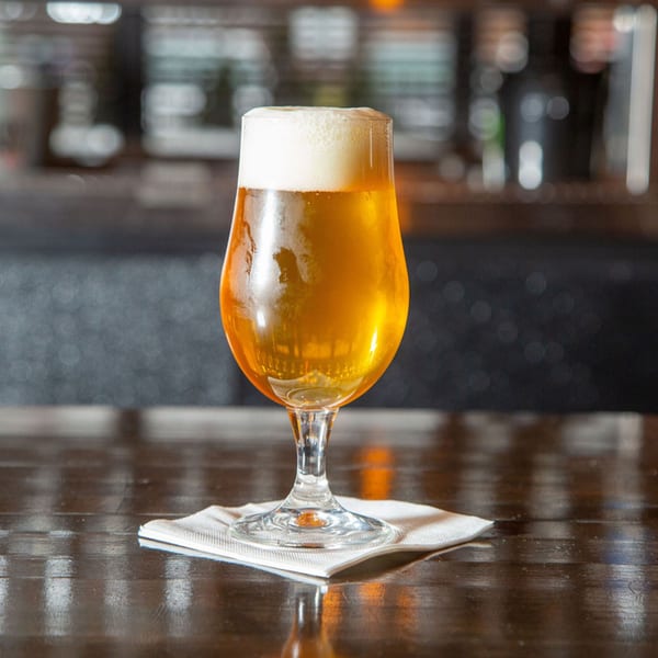 A glass of golden beer with a frothy head sits on a white napkin on a dark wooden bar.