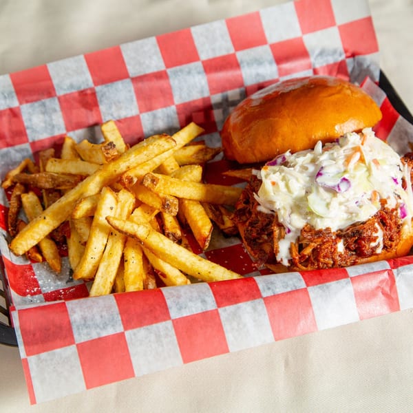 Pulled pork sandwich with coleslaw and a side of fries in a red and white checkered basket.