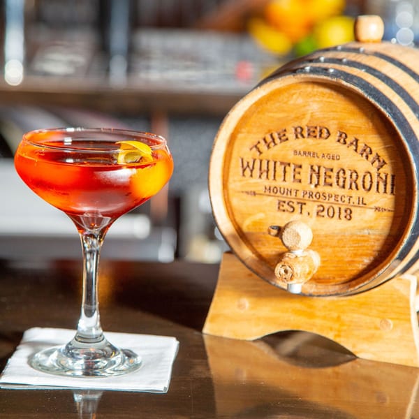 A Red Barn White Negroni cocktail in a coupe glass next to a branded barrel.